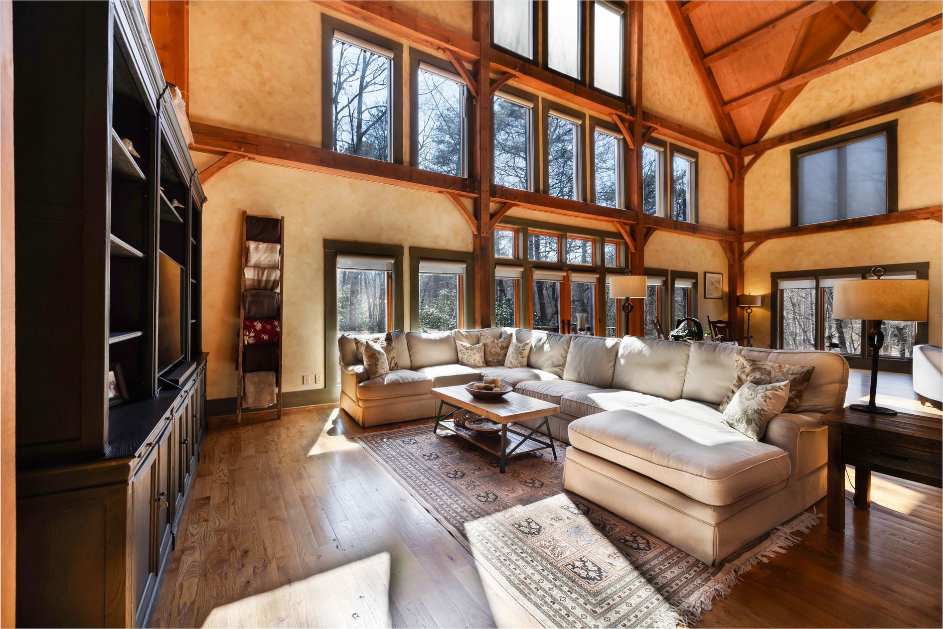 A bright living room with large windows, timber beams and pale seating, lit almost entirely by daylight.