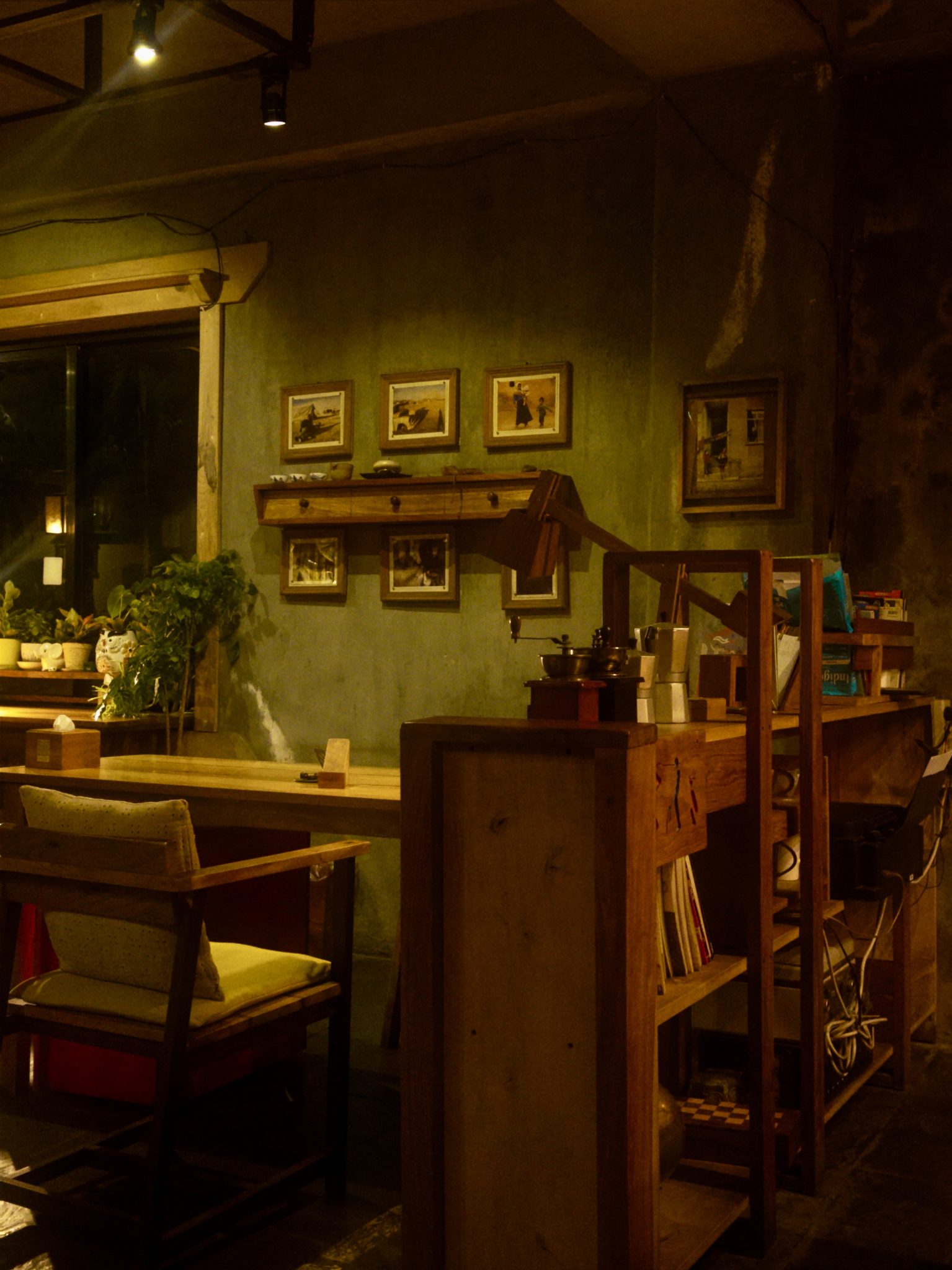 A dimly lit cafe interior with warm yellow lighting, wooden furniture, framed black and white photographs on a textured green wall, indoor plants on a windowsill, and a manual coff