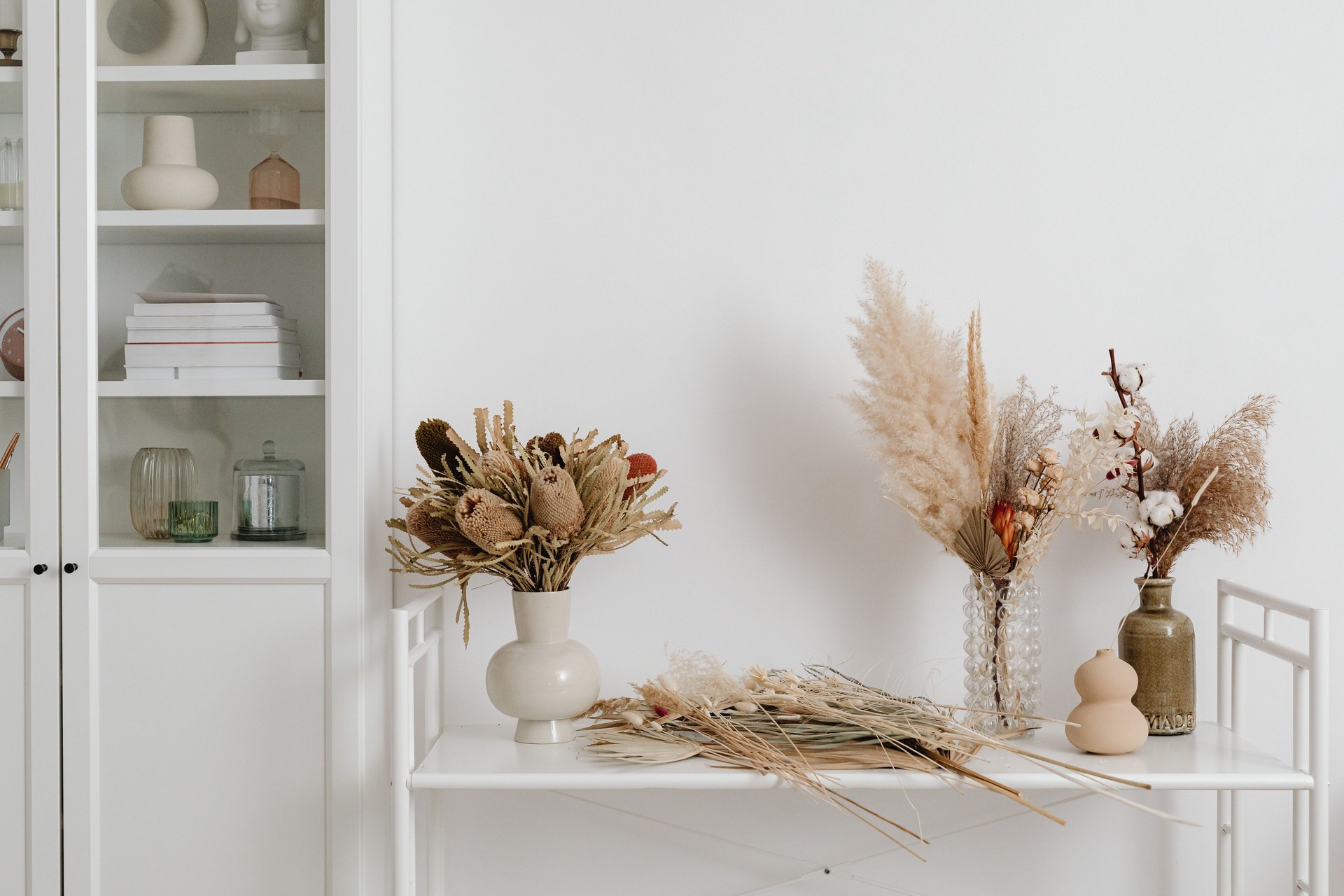 Dried flower arrangements in vases beside a white cabinet in a bright room
