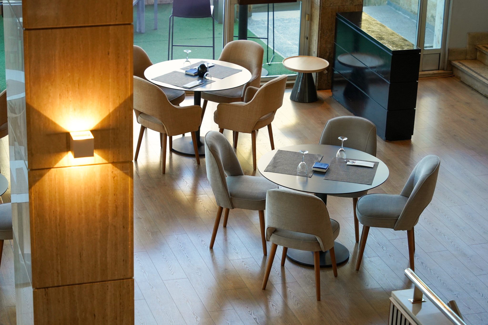 A modern, stylish restaurant interior with round tables surrounded by comfortable chairs. Tables are set with placemats, cutlery, and glassware. Natural light is streaming in from