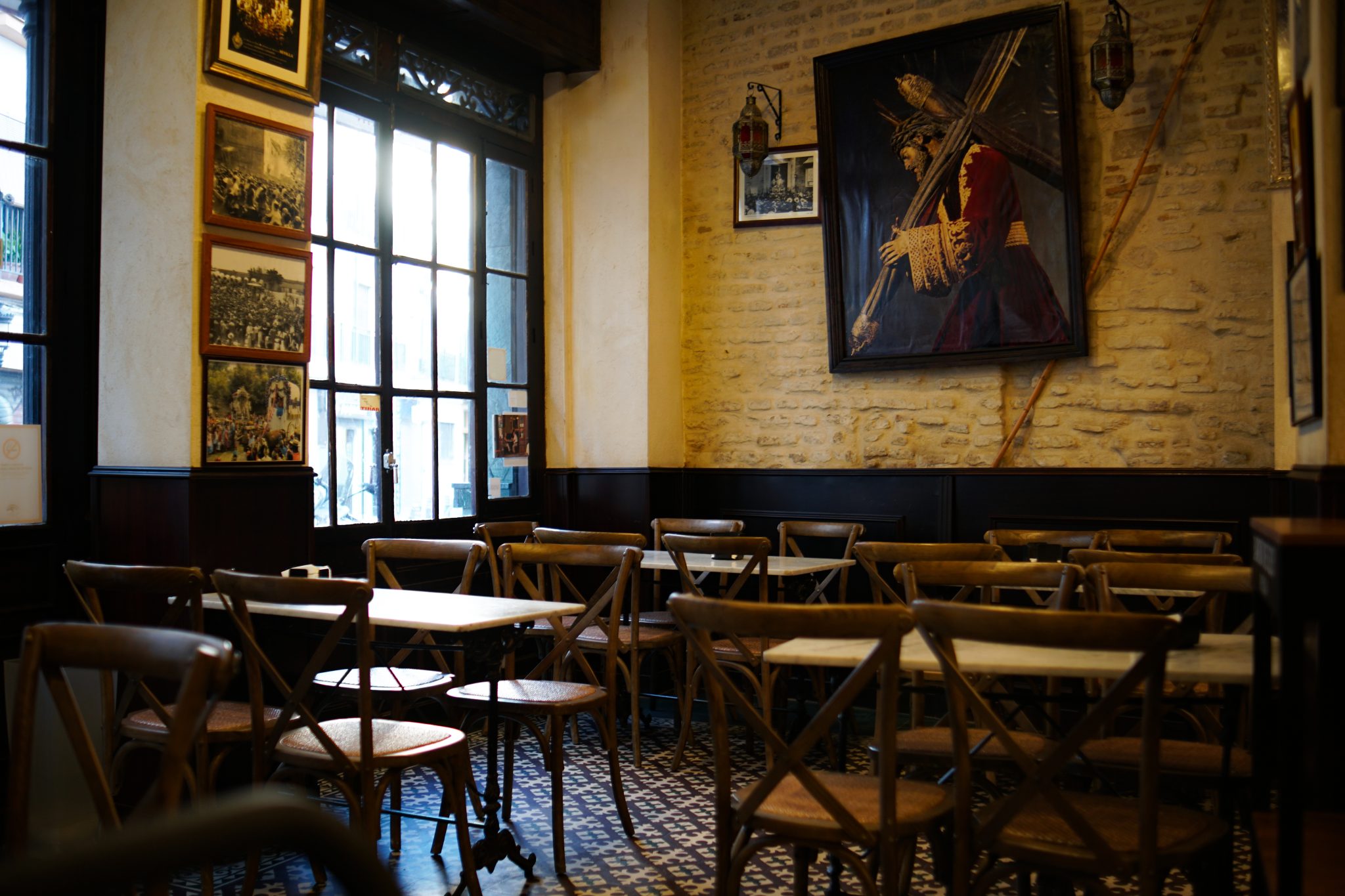 A cozy cafe interior with wooden chairs and tables arranged neatly on a patterned tile floor. Large windows allow natural light to illuminate the space. The decor features rustic e