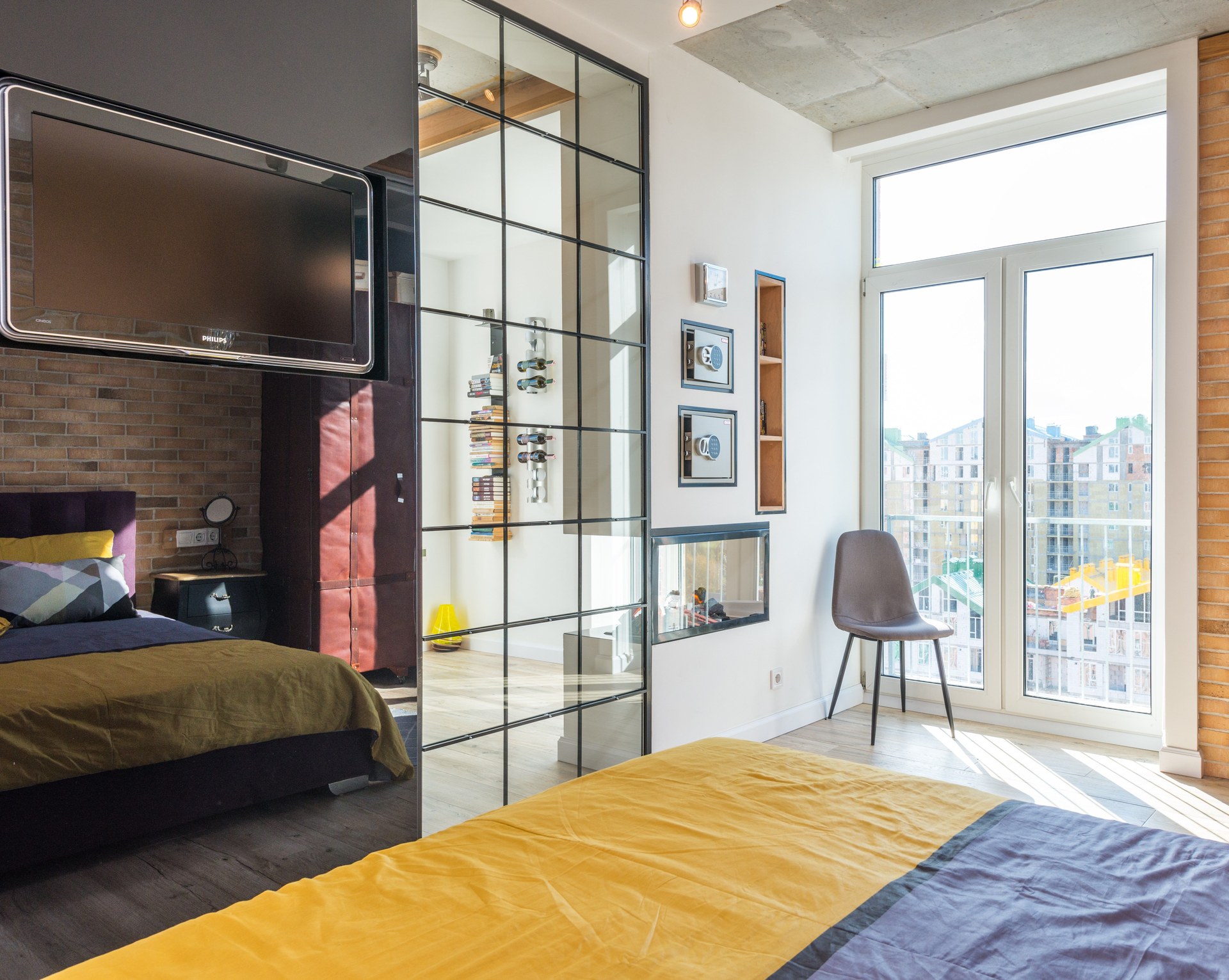 Comfortable bed with colorful bedclothes placed against black reflective wall with TV in light bedroom with decorations and glass door
