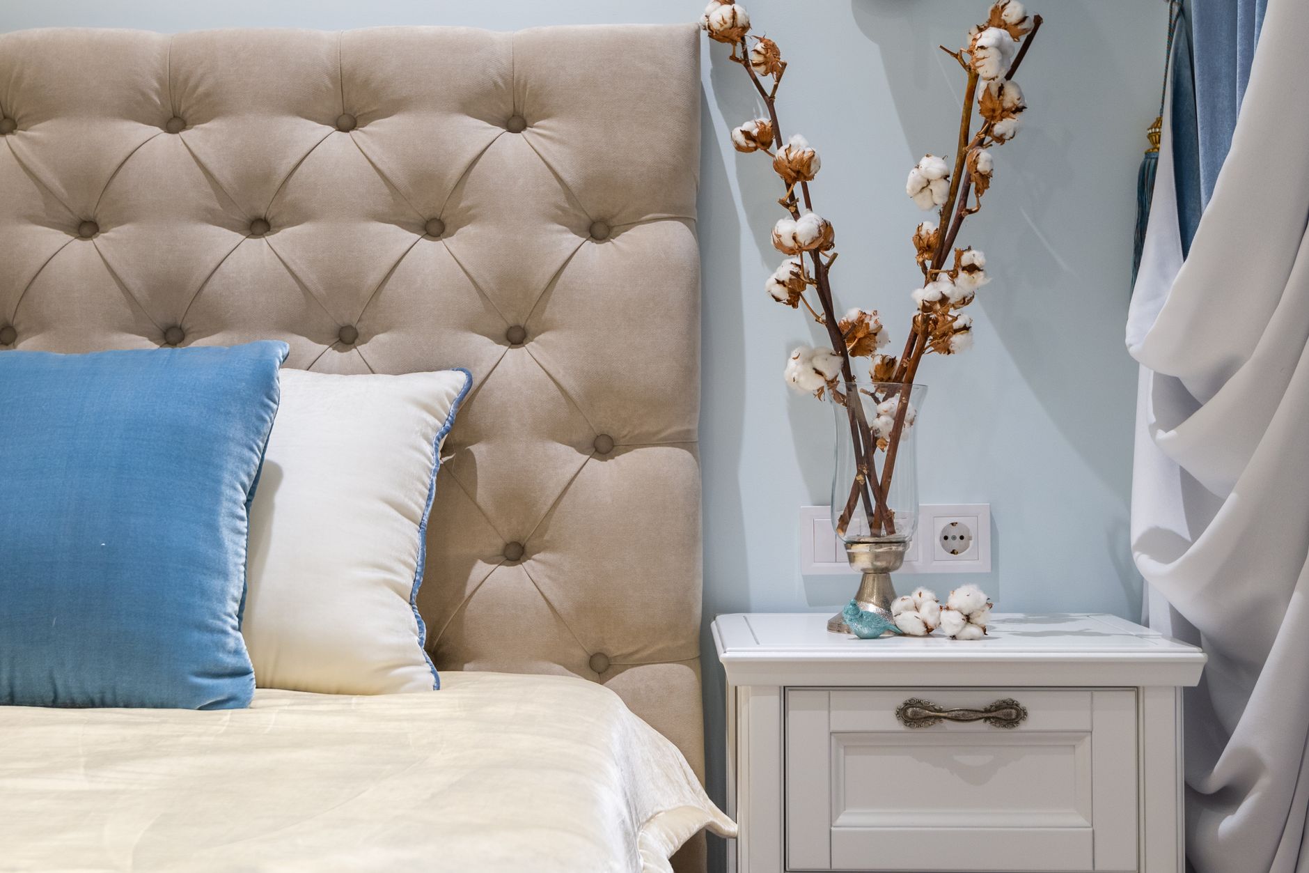 Cotton plants in vase placed on white nightstand near comfortable bed with soft cushions and headboard in classic styled bedroom, illustrating How Should a Headboard Be Positioned?
