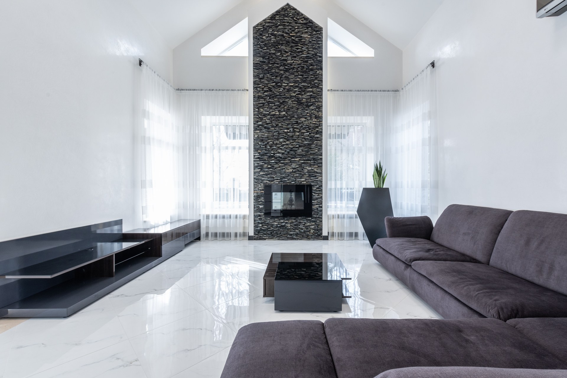 Big comfortable sofa and small table placed on shiny marble effect floor in cozy spacious living room with electric fireplace and green pott