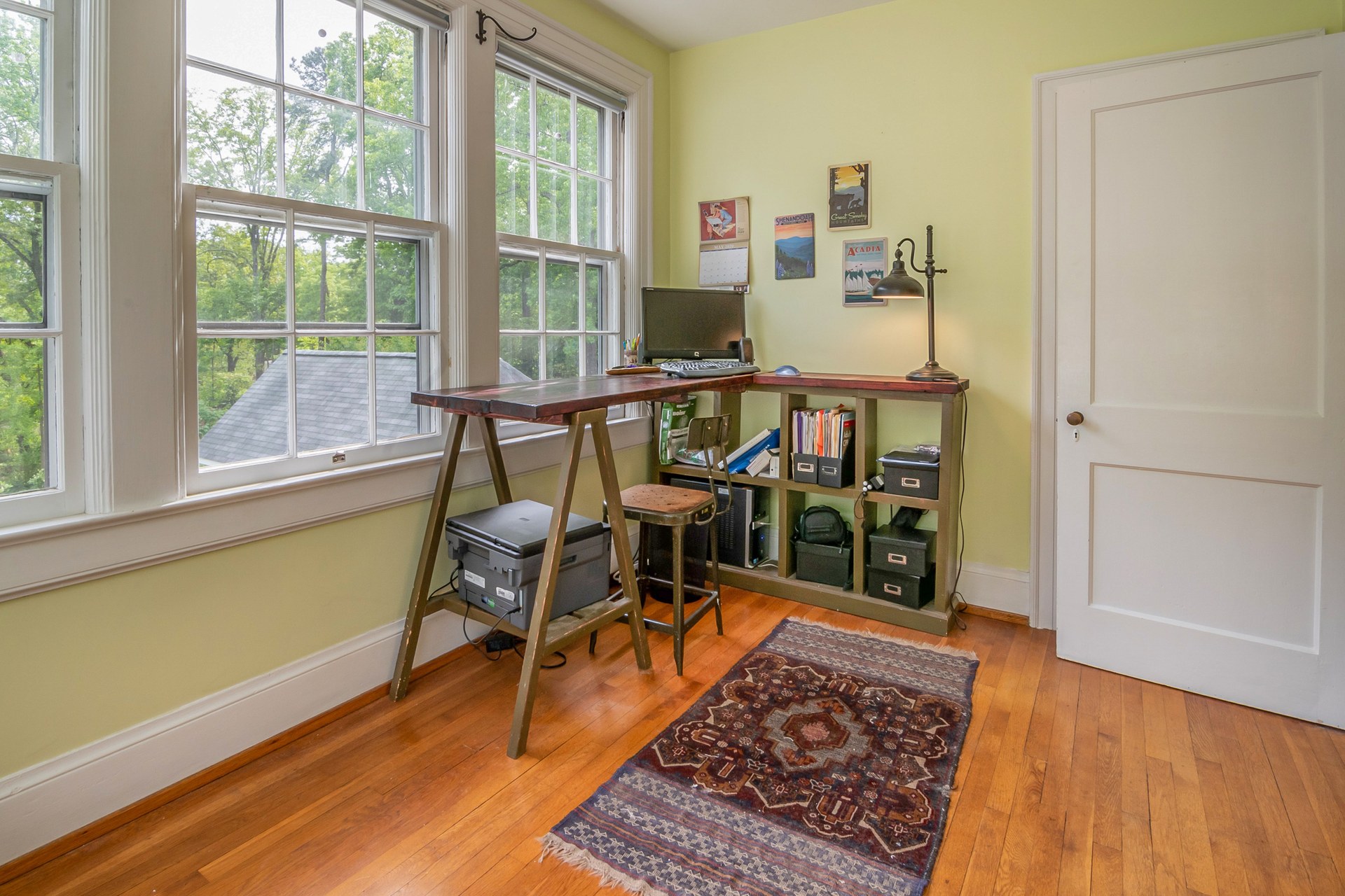 A wooden desk set into a corner beside tall windows in a home office