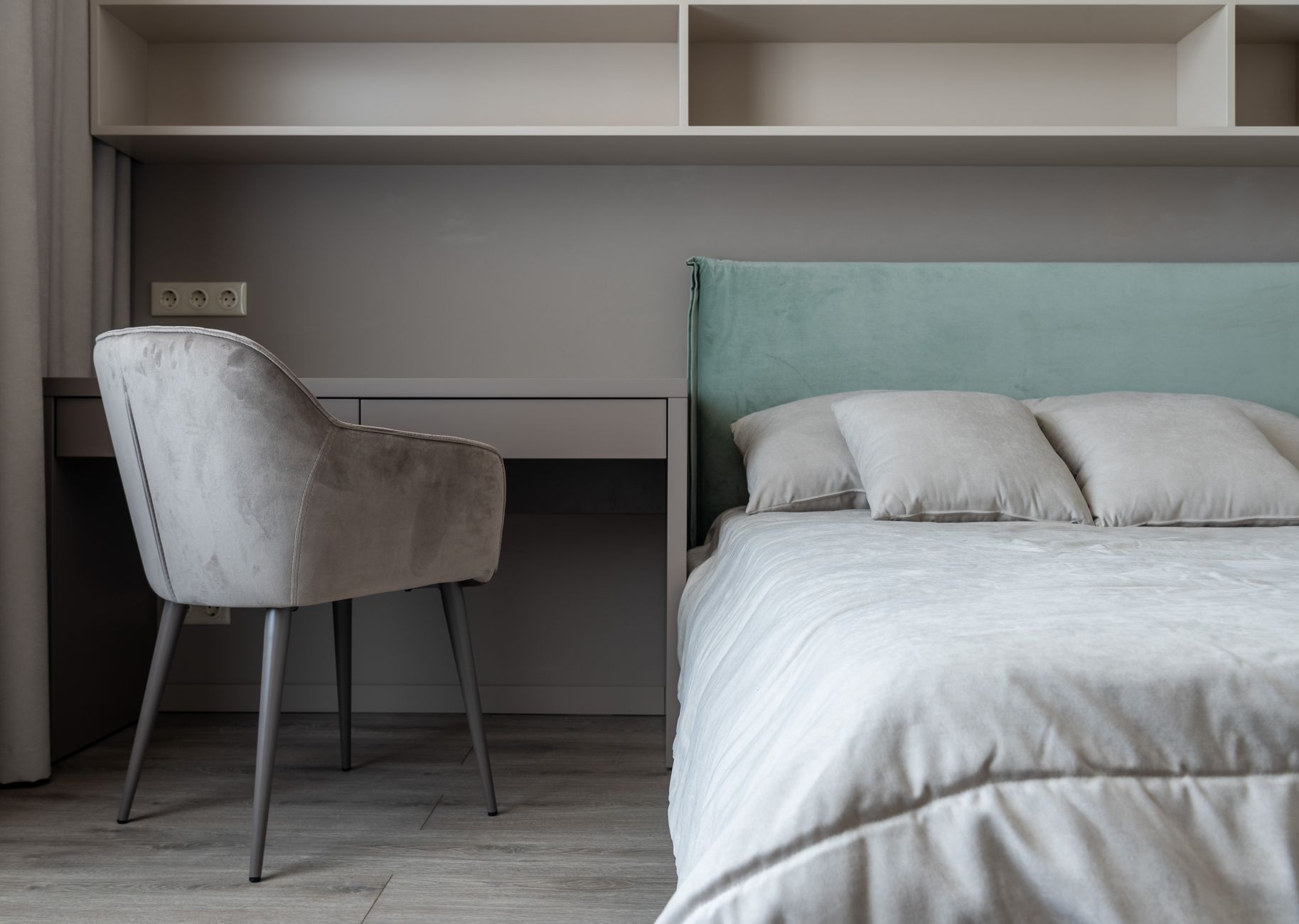 Interior of modern light bedroom with pillows and blanket on bed near table with chair under shelves next to curtains, illustrating Where Should the Bed Go in a Bedroom? A Feng Shu