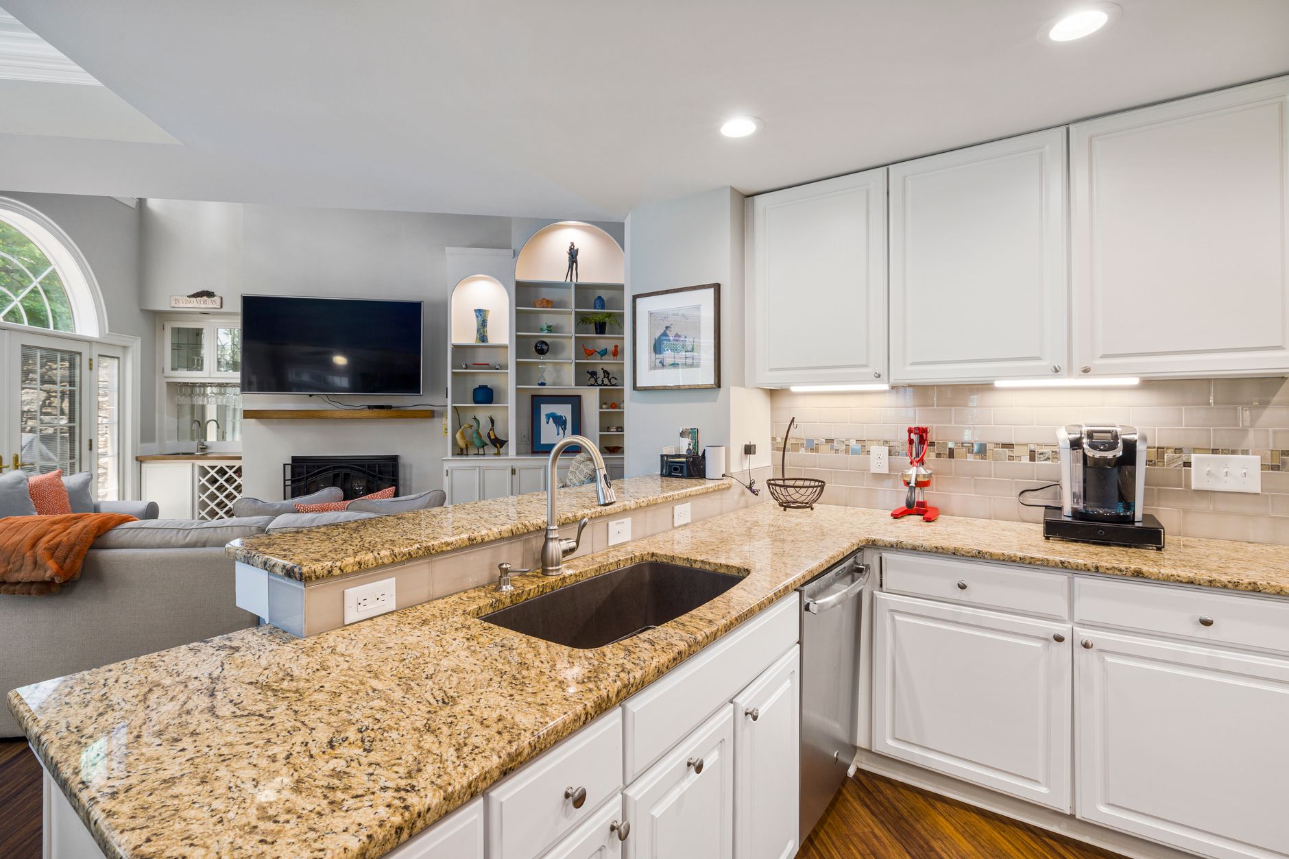 A modern kitchen with granite countertops opens into a stylish living room, featuring elegant furniture and built in shelves, illustrating Where Should the Kitchen Stove Be Placed?