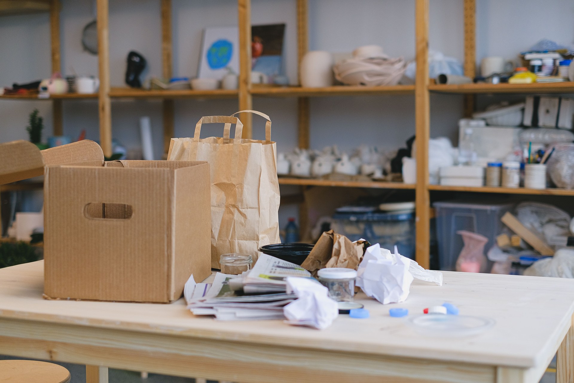 Cardboard box and paper bags on table in cluttered workspace with shelves. Ideal for packaging concepts